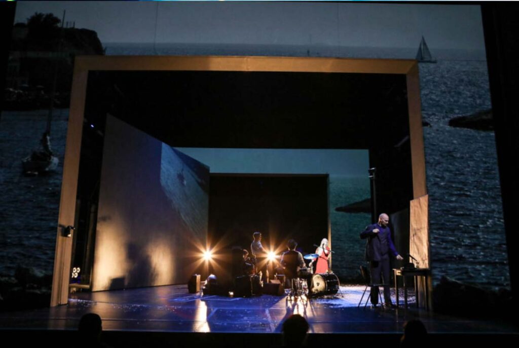 DER REISENDE,Theaterszene mit mehreren Darsteller:innen und Musikern in einer offenen Bühnenkonstruktion vor einer projizierten Meereslandschaft.