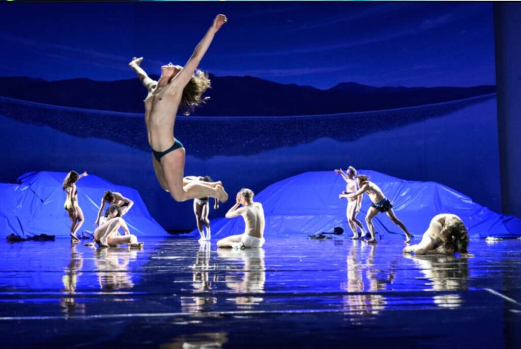 THE FUTURE, Volksbühne Berlin. Bühne: Alissa Kolbusch. Tänzer:innen auf einer blau beleuchteten Bühne mit reflektierendem Boden, eine Person springt hoch in die Luft.