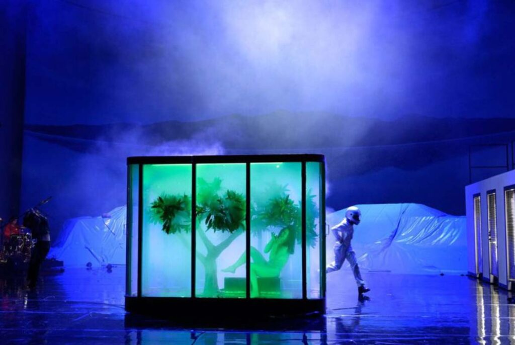 THE FUTURE, Volksbühne Berlin. Bühne: Alissa Kolbusch. Grün beleuchtete Glasbox mit Baum auf einer blau beleuchteten Theaterbühne, Tänzer:innen in der Box und im Hintergrund.