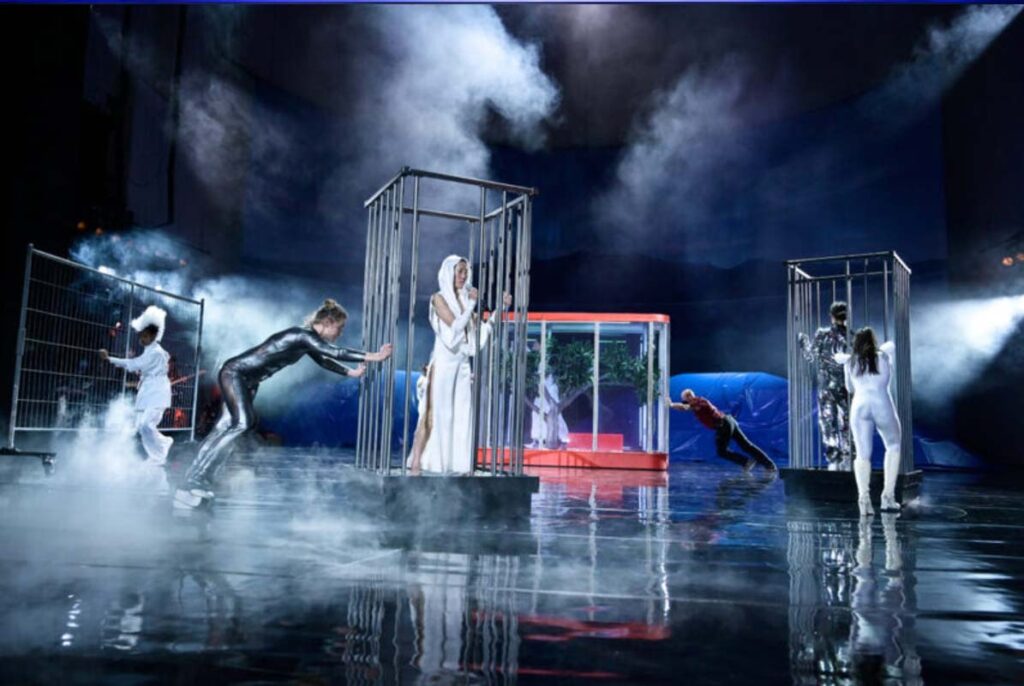 THE FUTURE, Volksbühne Berlin. Bühne: Alissa Kolbusch. Bühnenbild mit mehreren Figuren in Käfigen auf einer nebligen Theaterbühne,