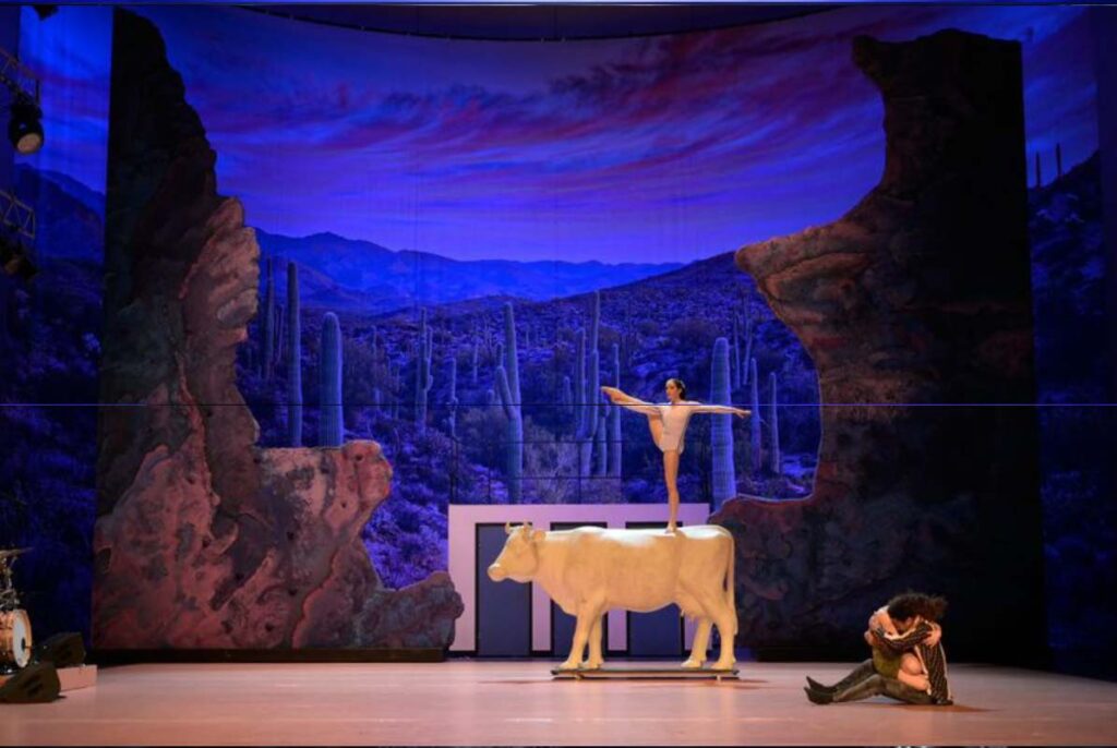 THE WEST, Volksbühne Berlin. Bühne: Alissa Kolbusch. Theaterbühne mit Wüstenlandschaft im Hintergrund; eine Person balanciert auf einer Kuhfigur.
