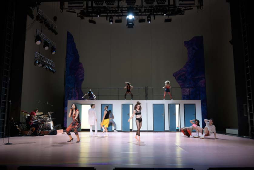 THE WEST, Volksbühne Berlin. Bühne: Alissa Kolbusch. Mehrere Darsteller:innen auf einer großen Theaterbühne mit erhöhtem Bühnenaufbau und Band.