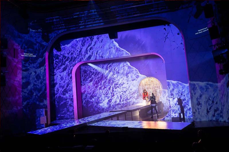 SLIPPERY SLOPE, Bühne: Alissa Kolbusch. Zwei Darsteller*innen hinten auf einer Bühne mit zwei überkreuzten Show-Stegen. Im Hintergrund drei runde Portale mit blau projiziertem Wasser