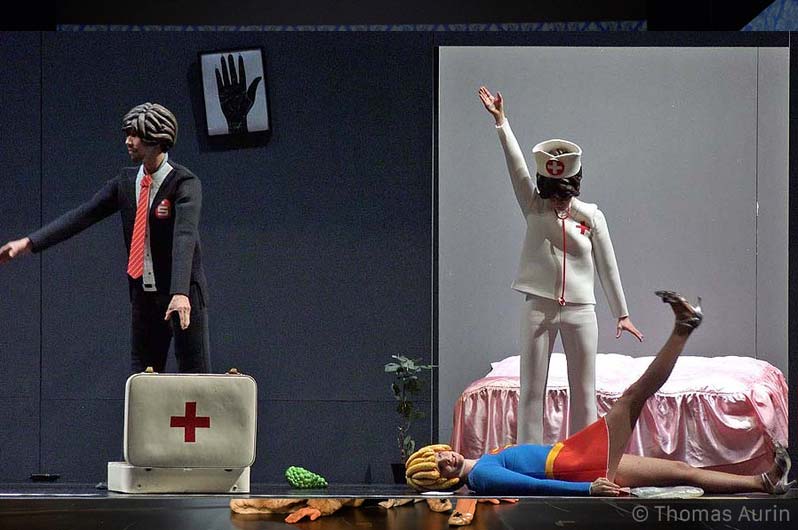 DER PALAST Volksbühne Berlin, Bühnenbild: Alissa Kolbusch. Drei Tänzer in Playmobil- Kostümen mit einem Notarztkoffer , eine liegt auf dem Boden, hinter ihr eine Playmobil- Krankenschwester und ein rosa Bett