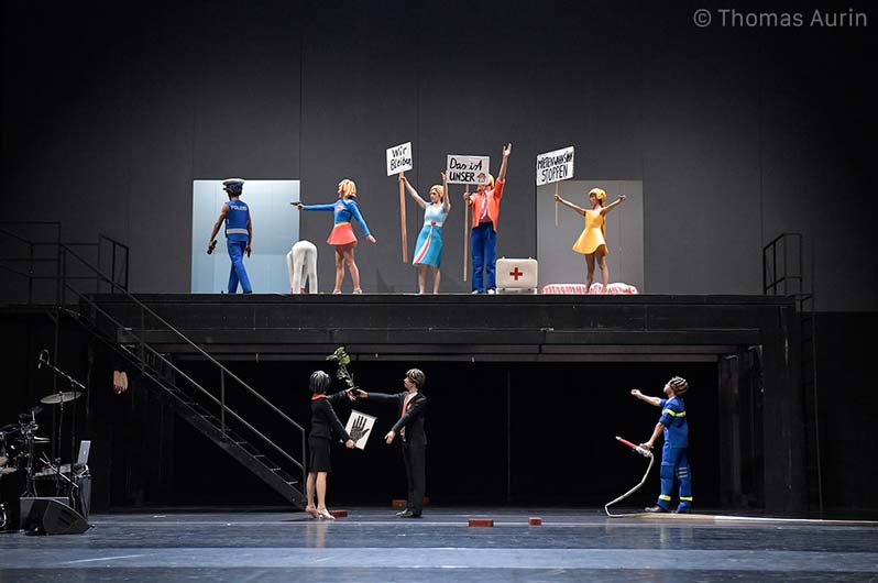 DER PALAST Volksbühne Berlin, Bühnenbild: Alissa Kolbusch Fünf Tänzer in Playmobil- Kostümen stehen auf einer Galerie und halten Schilder hoch, drei stehen unten vor einer Treppe