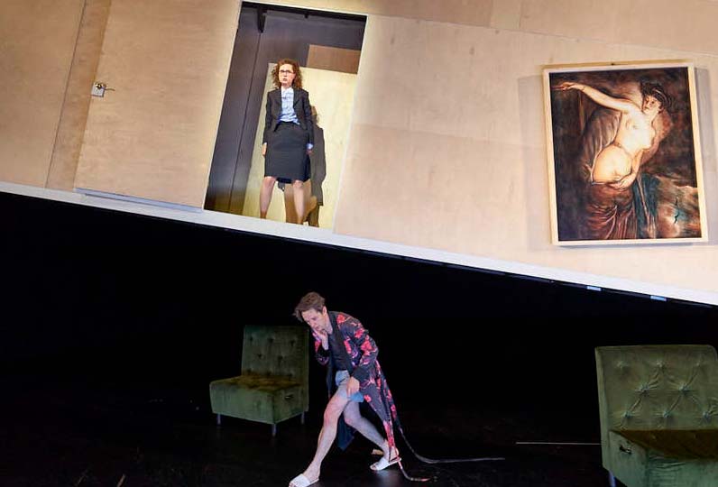 DER REVISOR Düsseldorfer Schauspielhaus, Bühne und Kostüme: Alissa Kolbusch. Eine Schauspielerin steht im Businesskostüm in der oberen Tür in der schräg hängenden Wand mit Bild, unten läuft der Schauspieler im Morgenmantel gekrümmt zwischen 2 Sitzbänken