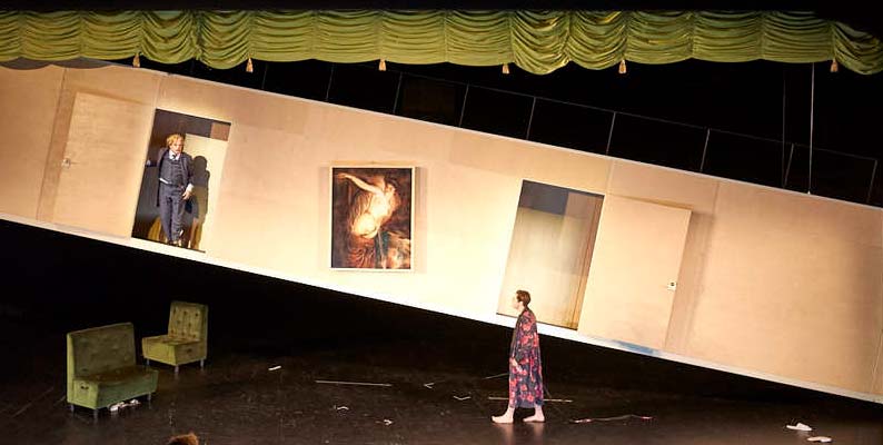 DER REVISOR Düsseldorfer Schauspielhaus, Bühne und Kostüme: Alissa Kolbusch. Eine schräg hängende Wand mit 2 Türen . Ein Schauspieler schaut aus der oberen Tür raus, der andere läuft im Morgenmantel unten auf dem Boden. Sitzbänke stehen herum und ein Bild hängt schief an der Wand. Oben ein grüner Raff- Vorhang.