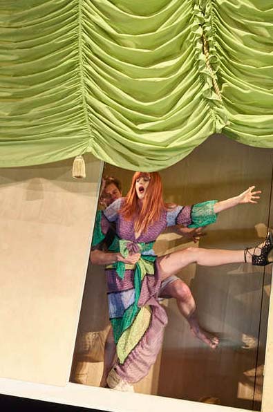 DER REVISOR Düsseldorfer Schauspielhaus, Bühne und Kostüme: Alissa Kolbusch. Ein Mann versucht eine rothaarige Frau( die Mutter ) in einem bunten Kleid von hinten zu umarmen , sie protestiert kokett. Oben ein grüner Raff- Vorhang.