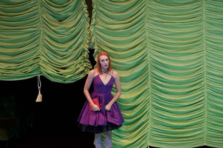 DER REVISOR Düsseldorfer Schauspielhaus, Bühne und Kostüme: Alissa Kolbusch. Eine junge Schauspielerin mit lila Kleid steht vor einem hellgrünen Raff- Vorhang
