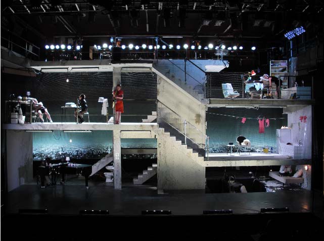 INSOMNIA Det kongelige Teater Kopenhagen, Bühne und Kostüme: Alissa Kolbusch. Ein großer mehrstöckiger Haus-Rohbau mit einem Treppenturm und fünf Zimmern. In der Küche kinks oben schläft ein Mann auf dem Stuhl, eine GFrau sitzt am Küchentisch. Unter ihnen sitzt ein Pianist am Flügel, rechts oben schläft eine Frau im Stehen in ihrem Büro.