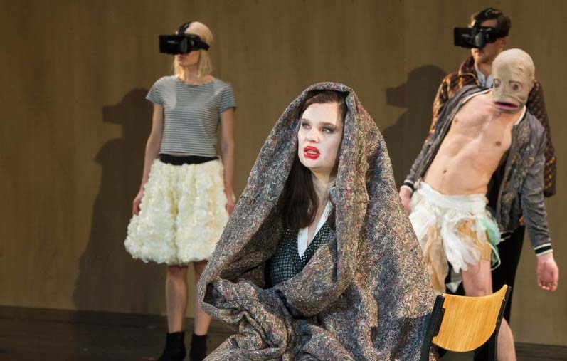 DER STURM Theater Bonn, Kostüme: Alissa Kolbusch. Eine Frau und ein Mann stehen hinten mit 3D Masken , eine Frau sitzt vorne in eine Decke gewickelt, hinter ihr Schauspiler mit einem Fischkopf.