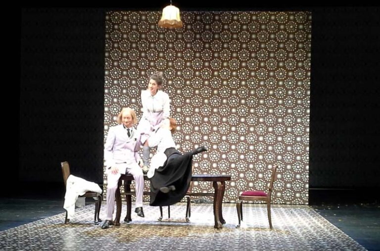 STERNHEIM TRILOGIE Staatstheater Braunschweig, Bühne: Alissa Kolbusch. Eine Schauspielerin steht, zwei Schauspieler*innen sitzen auf einem Esstisch, hinten eine Wand mit Tapeten.