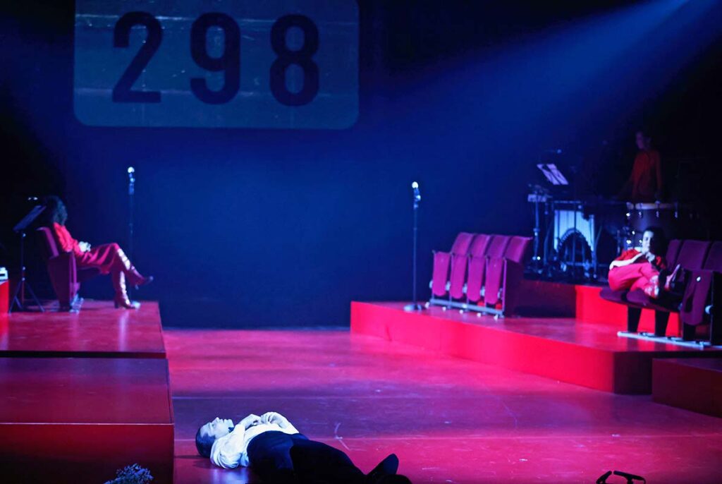 VATERMAL, Theaterszene mit roter Bühne, mehreren Darsteller:innen auf Sitzplätzen und einer liegenden Person im Vordergrund, während im Hintergrund die Zahl „298“ projiziert wird.