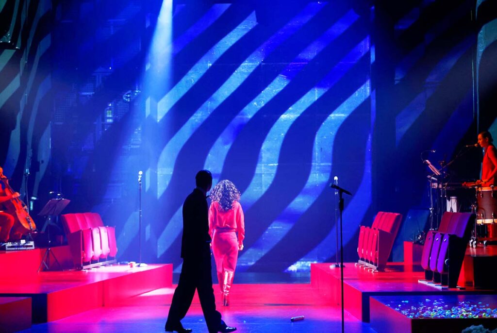 VATERMAL, Theaterszene mit blauem Licht und grafischem Hintergrund, während eine Darstellerin in rotem Kostüm über die Bühne geht und Musiker seitlich spielen.