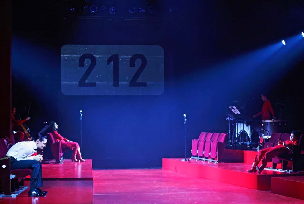 VATERMAL, Theaterszene mit roten Bühnenpodesten und Kinositzen in denen mehreren Darsteller:innen sitzen, sowie Musikern, während im Hintergrund die Zahl „212“ projiziert wird.