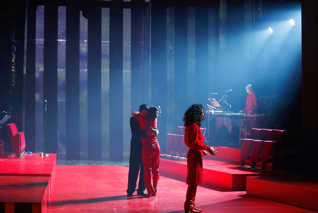 VATERMAL, Theaterszene mit drei Personen in roten Kostümen auf einer roten Bühne, während eine Musikerin im Hintergrund spielt.