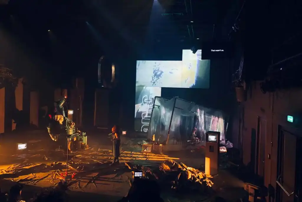 GROUND CONTROL Maxim Gorki Theater Berlin, Bühne: Alissa Kolbusch. Eine Darstellerin steht in der Mitte der Bühne iund spricht mit der Darstellerin, die auf dem Roboterarm sitzt. Auf dem Boden und im Hintergrund eine Videoinstallation in orangenem Licht.