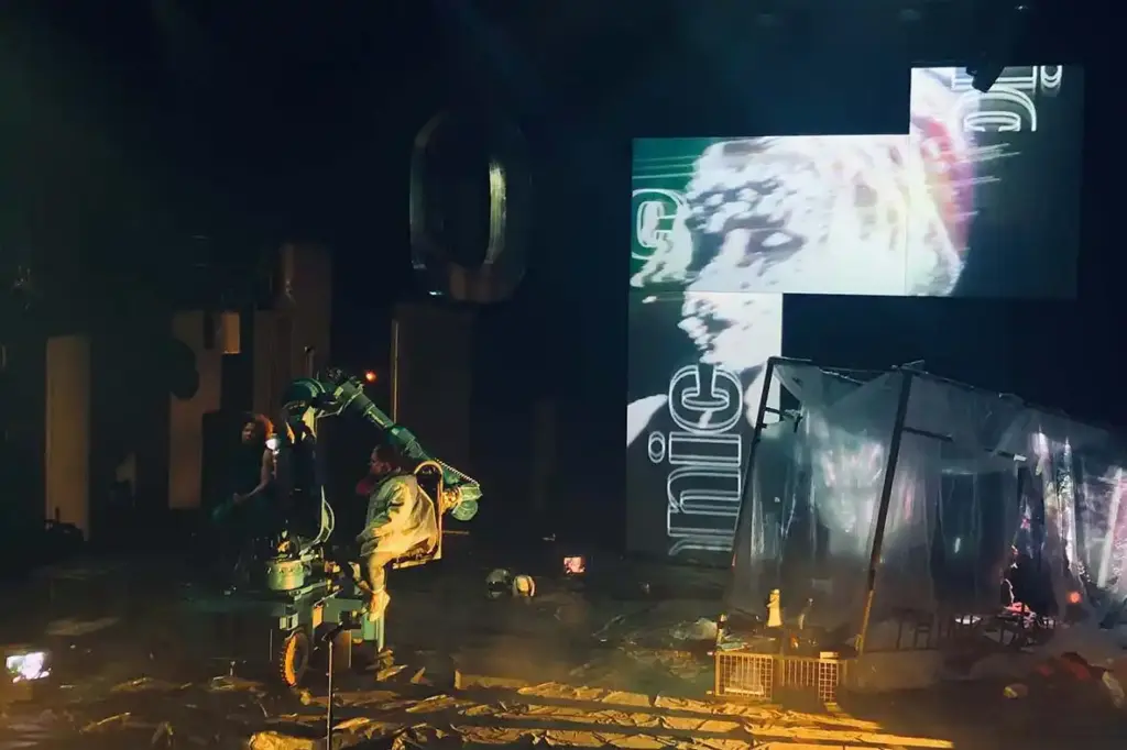 GROUND CONTROL Maxim Gorki Theater Berlin, Bühne: Alissa Kolbusch. Eine Darstellerin steht in der Mitte der Bühne iund spricht mit der Darstellerin, die auf dem Roboterarm sitzt. Auf dem Boden und im Hintergrund eine Videoinstallation in orangenem Licht.