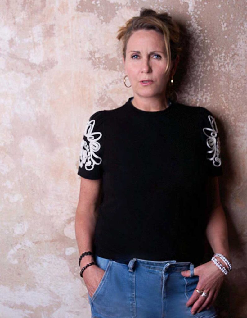Alissa Kolbusch, steht vor einer beigefarbenen, strukturierten Wand und trägt ein schwarzes T-Shirt mit weißen Mustern an den Ärmeln sowie eine blaue Jeans.