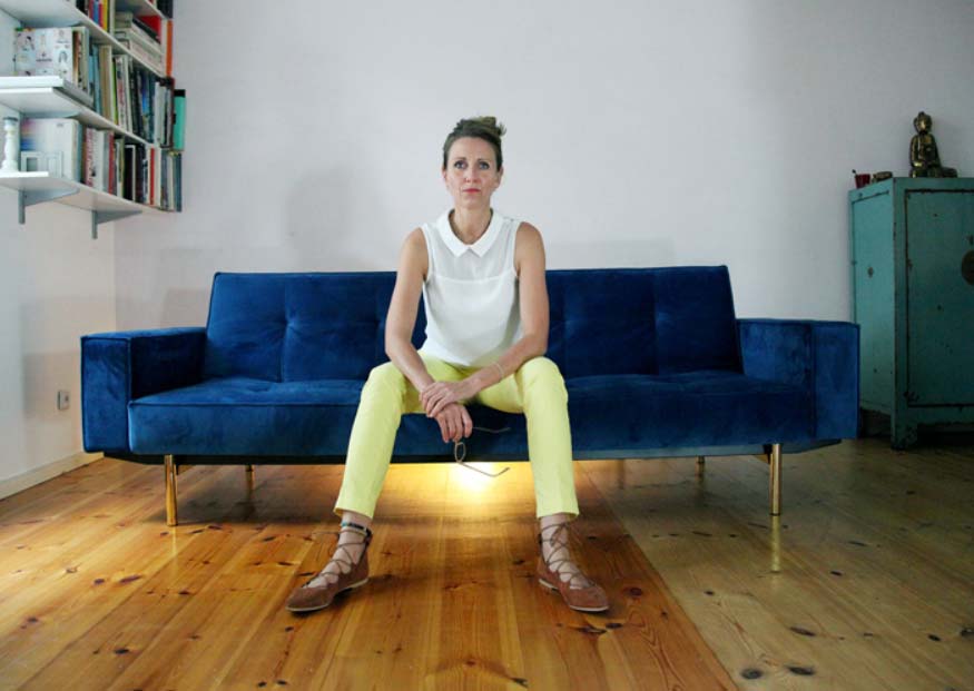 Alissa Kolbusch, Frau sitzt auf einem blauen Sofa in einem hellen Raum mit Holzboden. Sie trägt ein weißes Oberteil, gelbe Hose und Sandalen und blickt nach vorne. Links steht ein Bücherregal, rechts ein grüner Schrank.